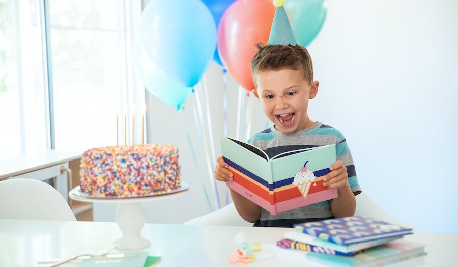 Little boy looking at a Chatbook with balloon and a cake around him.